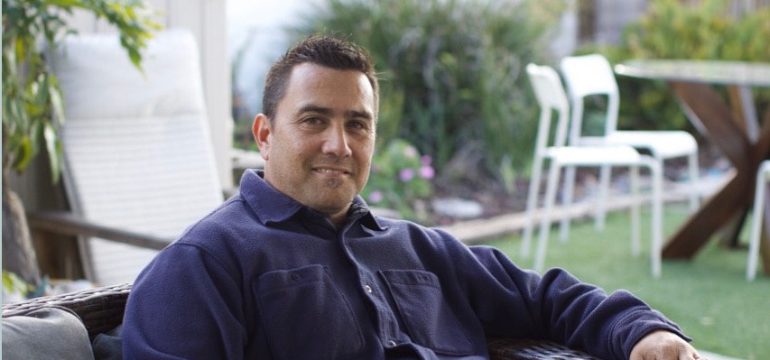 LeifQ is sitting comfortably on a patio chair, smiling at the camera, with greenery and outdoor furniture in the background.